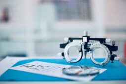 A pair of adjustable trial frames rests on a desk beside an eye chart and a pair of lenses in an optometry clinic.