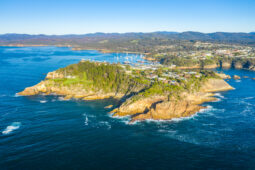 Aerial view of a rocky coastal headland with a small town, surrounded by bright blue ocean and distant forest-covered hills.