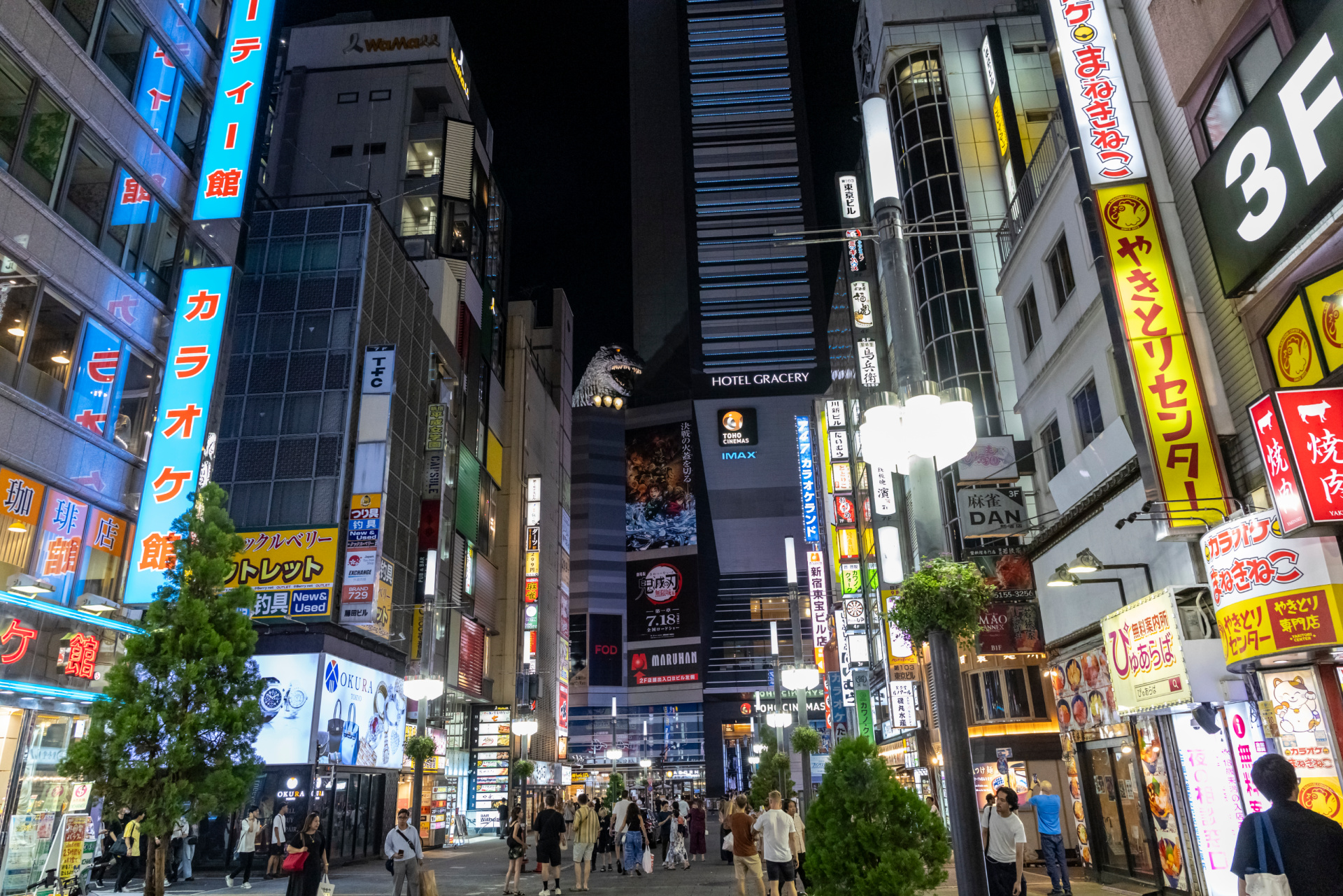 A busy Tokyo street at night, lined with tall buildings covered in bright neon signs and billboards, with crowds of people walking beneath the lights.