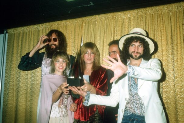 Five musicians from Fleetwood Mac stand together on stage holding a trophy, dressed in 1970s-style clothing, with one person in the foreground extending a hand toward the camera.