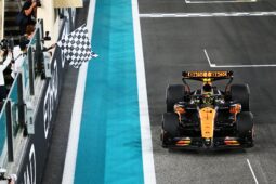 A Formula 1 car driven by Lando Norris crosses the finish line as a marshal waves the chequered flag from the pit wall.