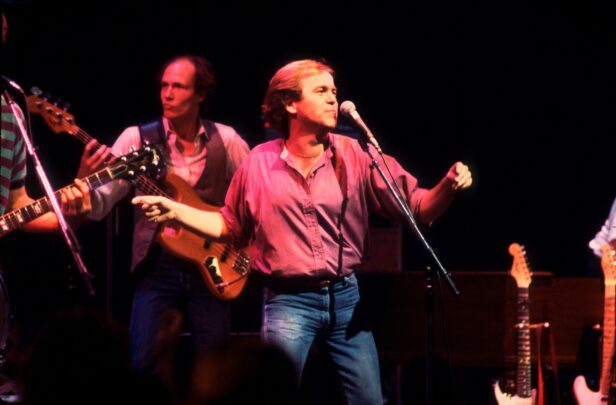 A male singer performs on stage at a live concert, standing at a microphone with arms slightly outstretched while band members play behind him.