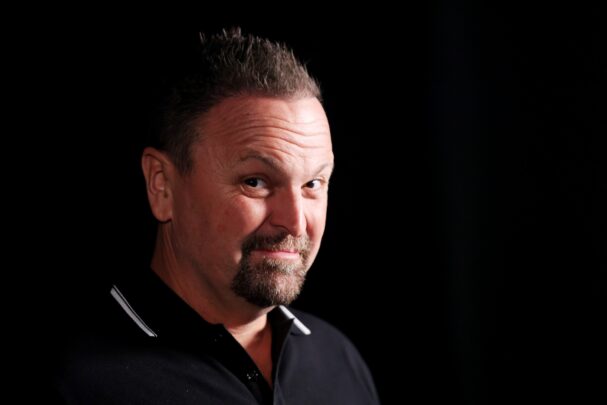 Middle-aged man with short dark hair and a trimmed goatee looks toward the camera with a knowing half-smile against a black background.