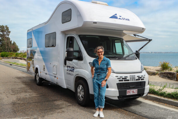 A smiling woman stands beside a white campervan parked near the water on a coastal road.