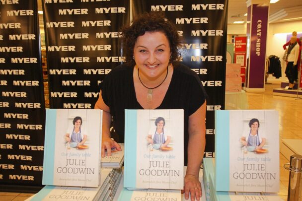 A smiling author stands behind a table displaying copies of her cookbook at a department store signing event.