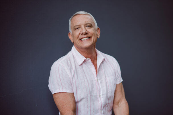 A smiling man in a light pink shirt poses against a dark background.