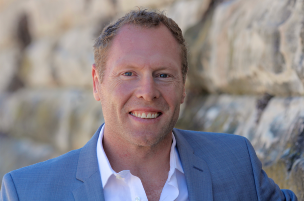 Portrait of a smiling man in a blue suit standing outdoors against a rocky background.
