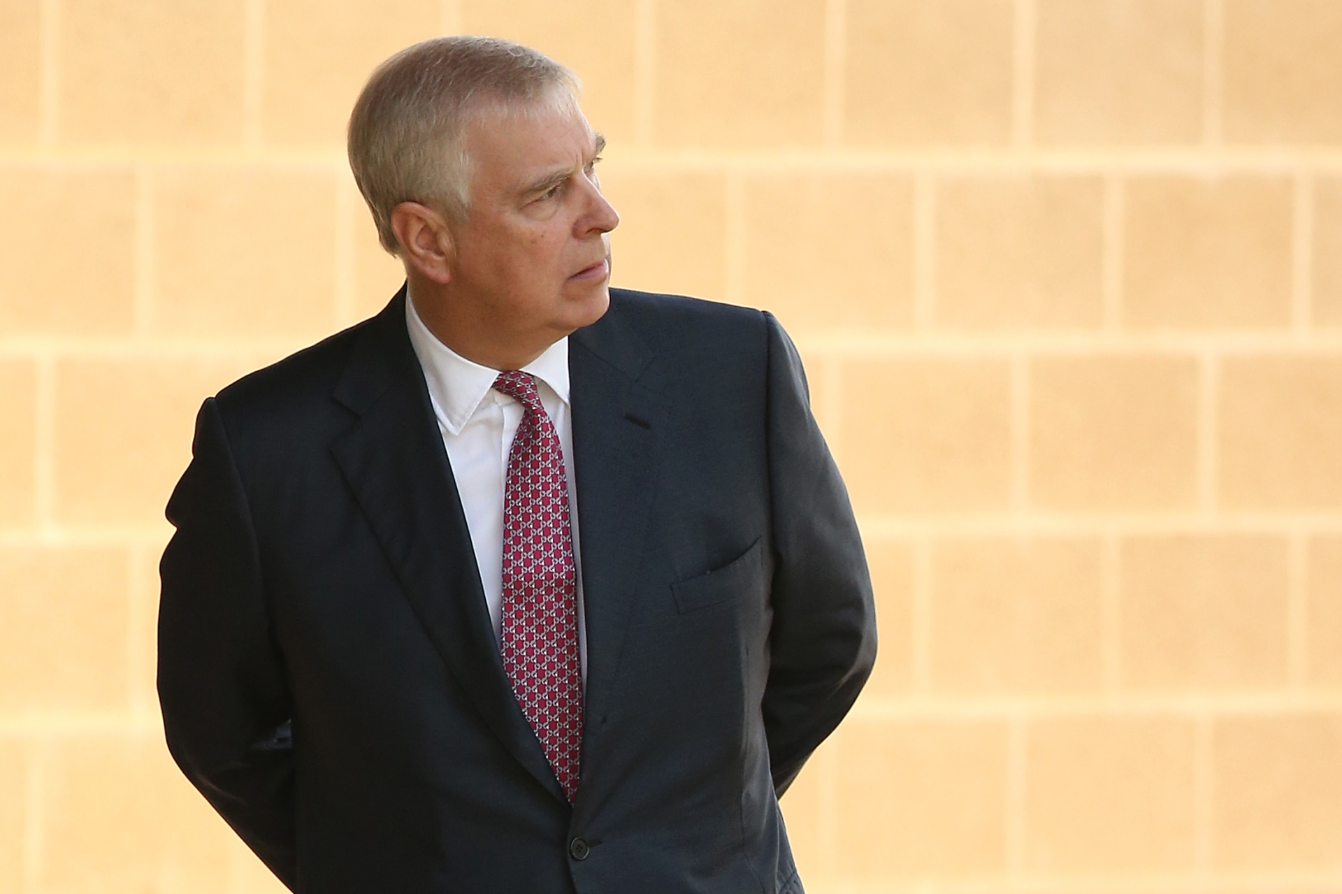 A man in a dark suit and red patterned tie stands with his hands behind his back, looking to the side.