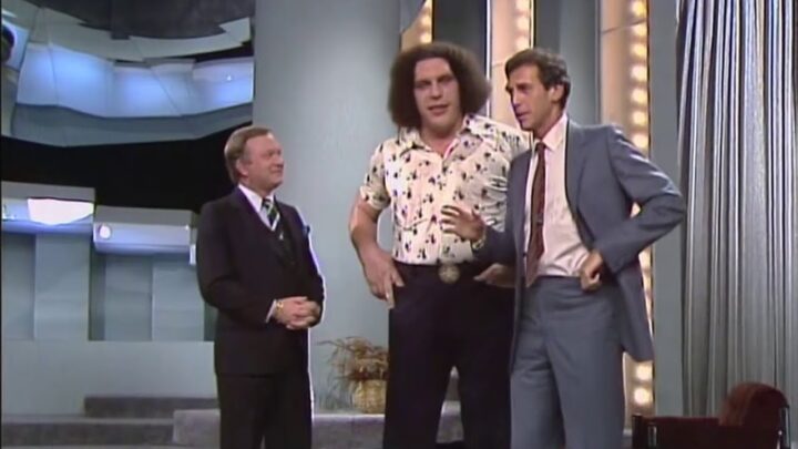 Three men stand on a brightly lit television studio stage during a talk show segment.