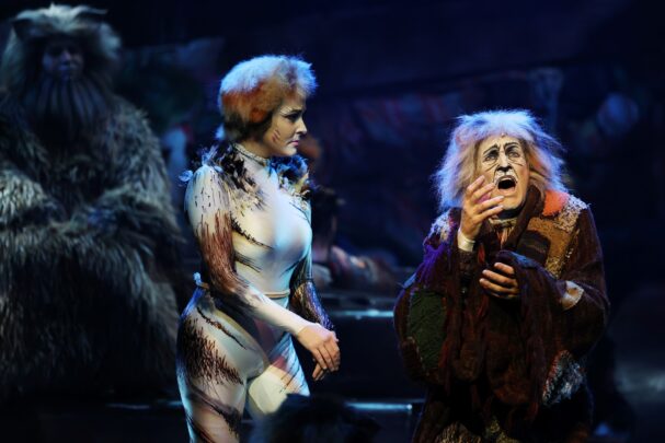 Two performers in theatrical cat costumes sing on stage under blue lighting.