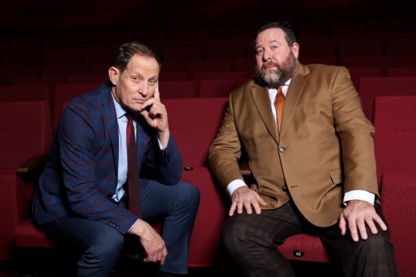 Two men in suits sit in red theatre seats, posing for a promotional photo.