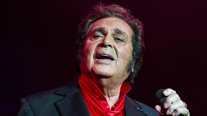 Engelbert Humperdinck singing on stage while holding a microphone under red lighting.
