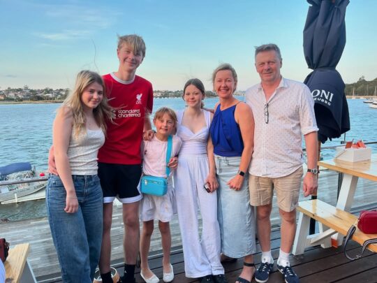 Family of six smiling and posing together on a waterfront deck with boats and houses in the background.