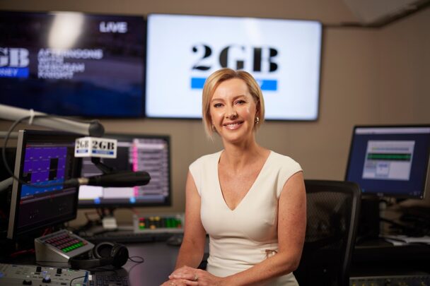 Woman seated in a radio studio with microphones and computer screens displaying station branding.