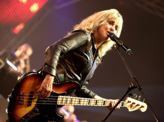 Suzi Quatro singing into a microphone while playing bass guitar on stage.